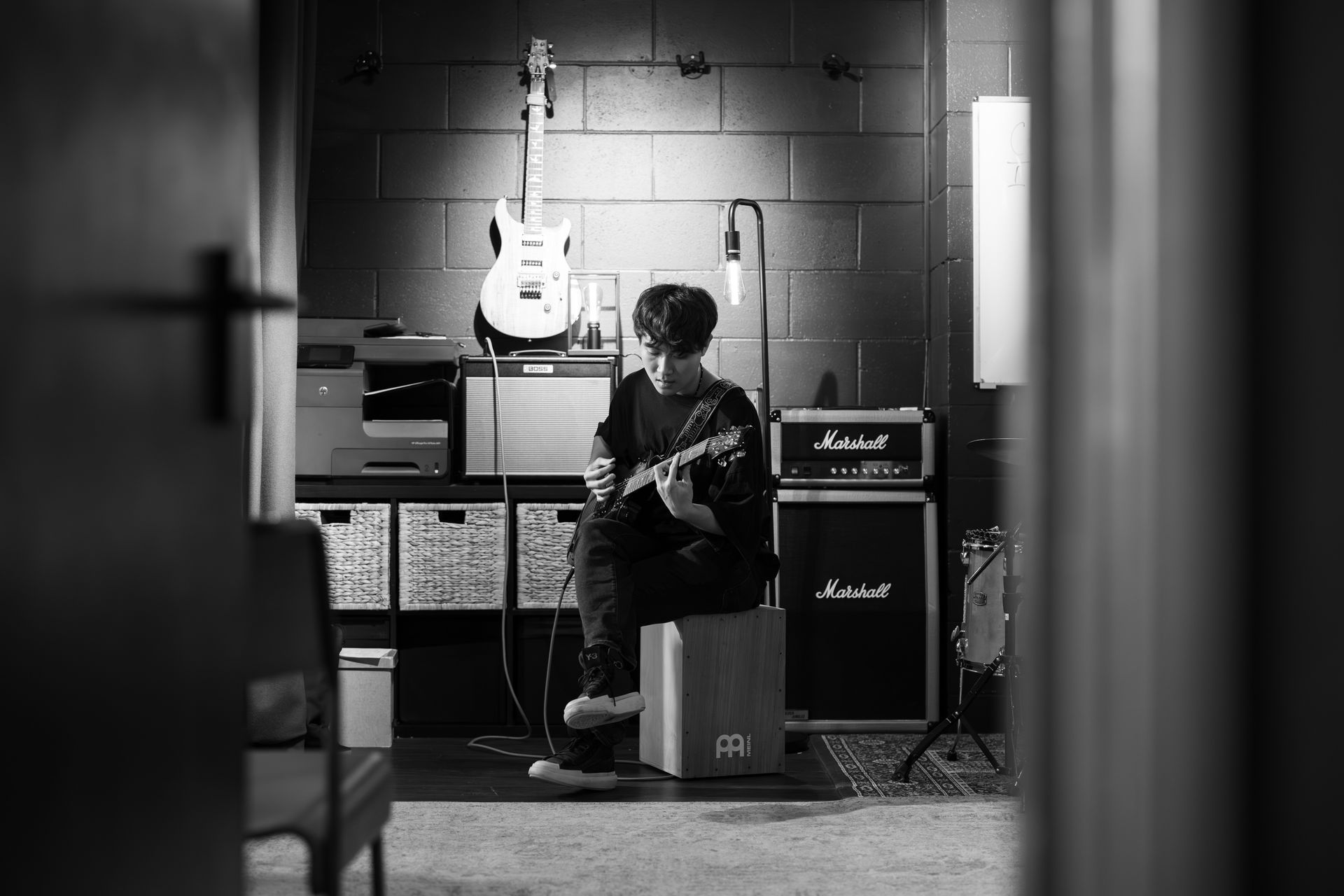 Person playing guitar in a studio, facing away. Guitars, amps, and recording equipment are in the background. Black and white.