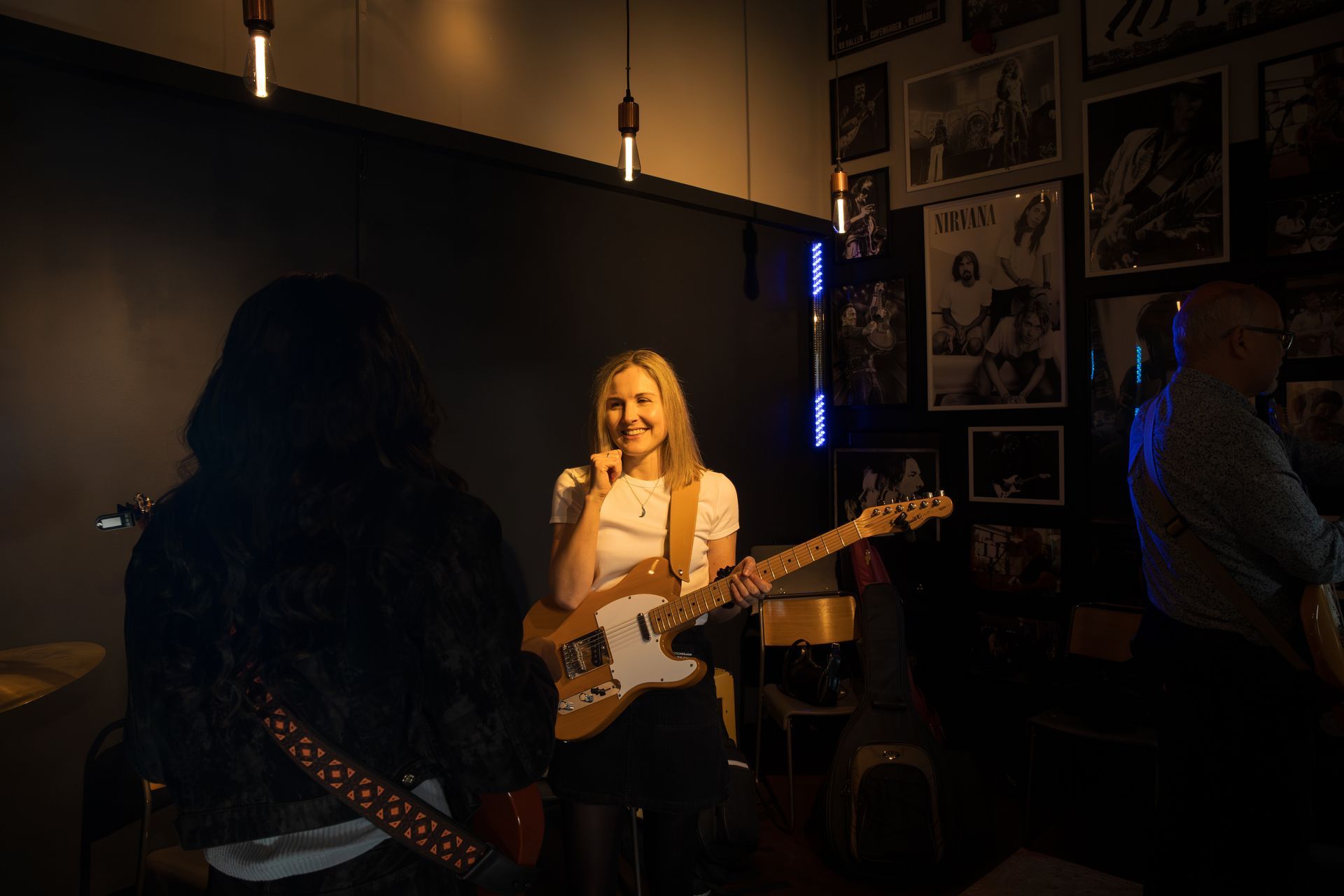 Band performing. Blonde woman with guitar smiles, standing center stage. Dark room, posters on wall.