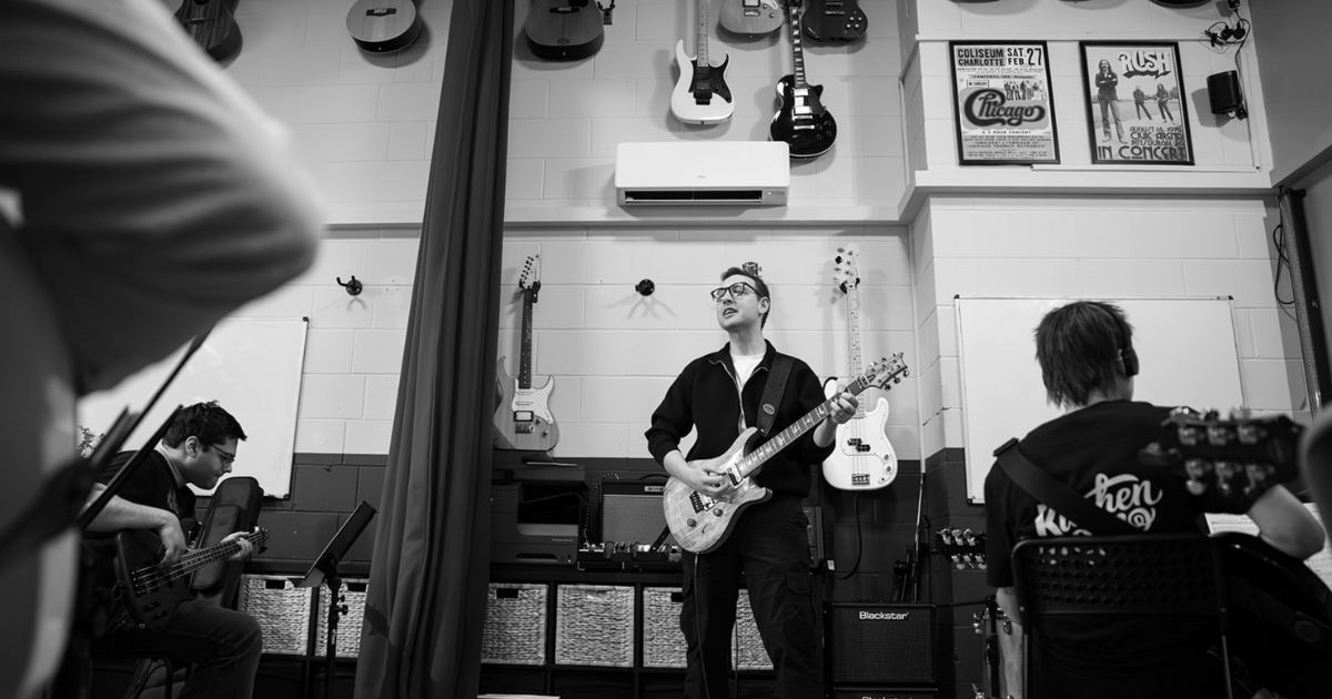 Band rehearsing in a room with guitars on the wall. A guitarist plays center stage.