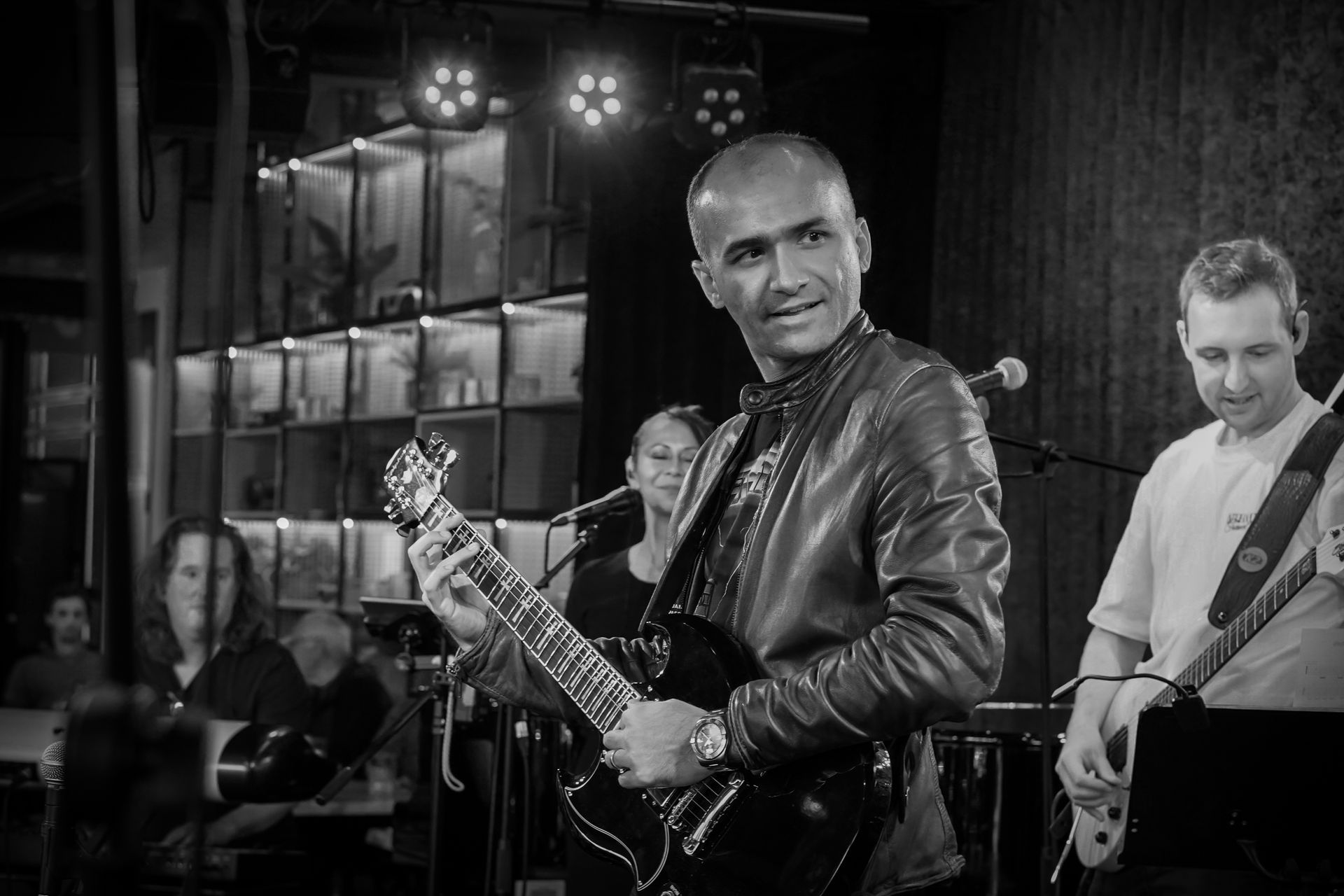 Band performing on stage, guitarist in leather jacket, other musicians in background.