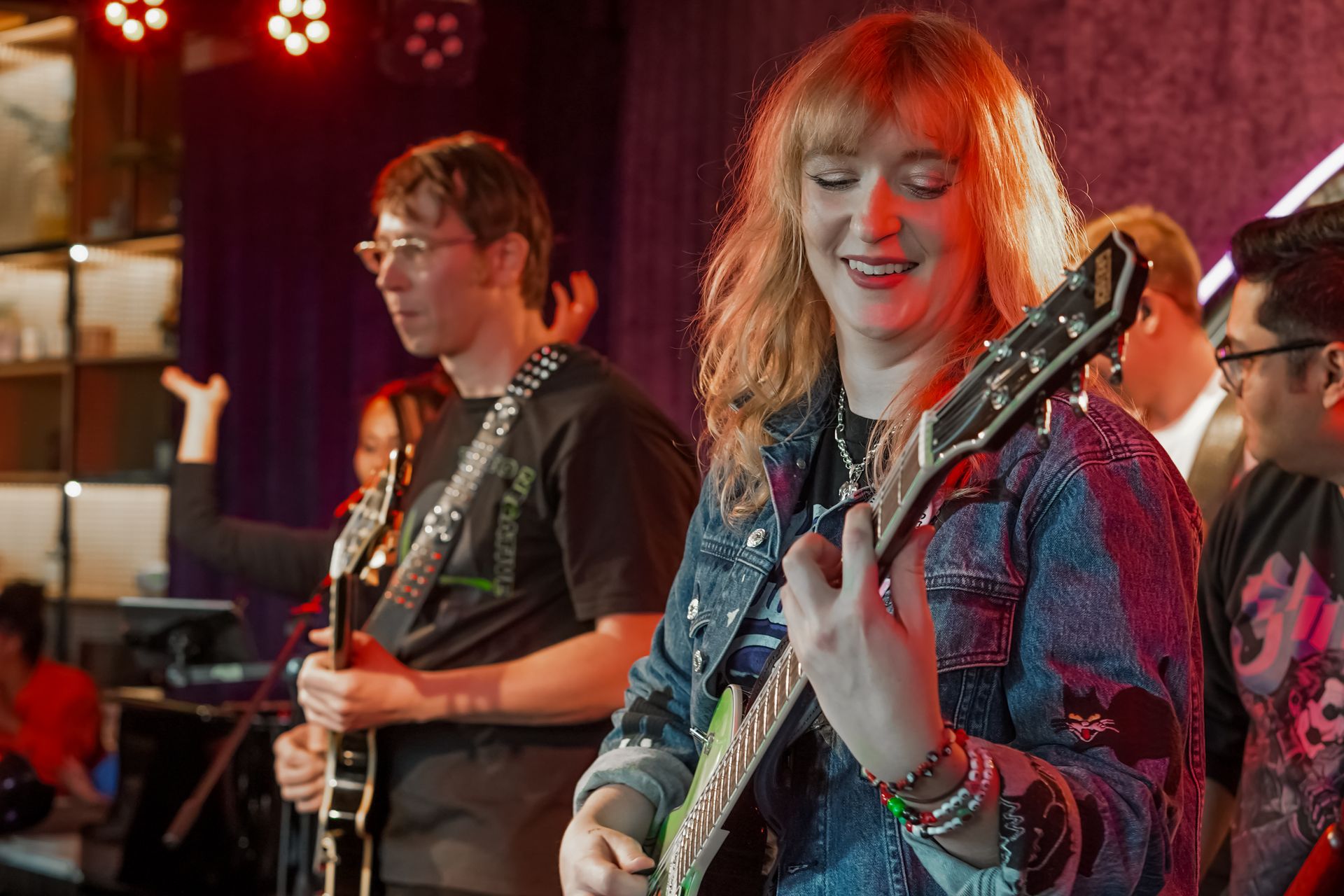 Band performing on stage, guitarist in denim jacket smiles, others play instruments with dim lighting.