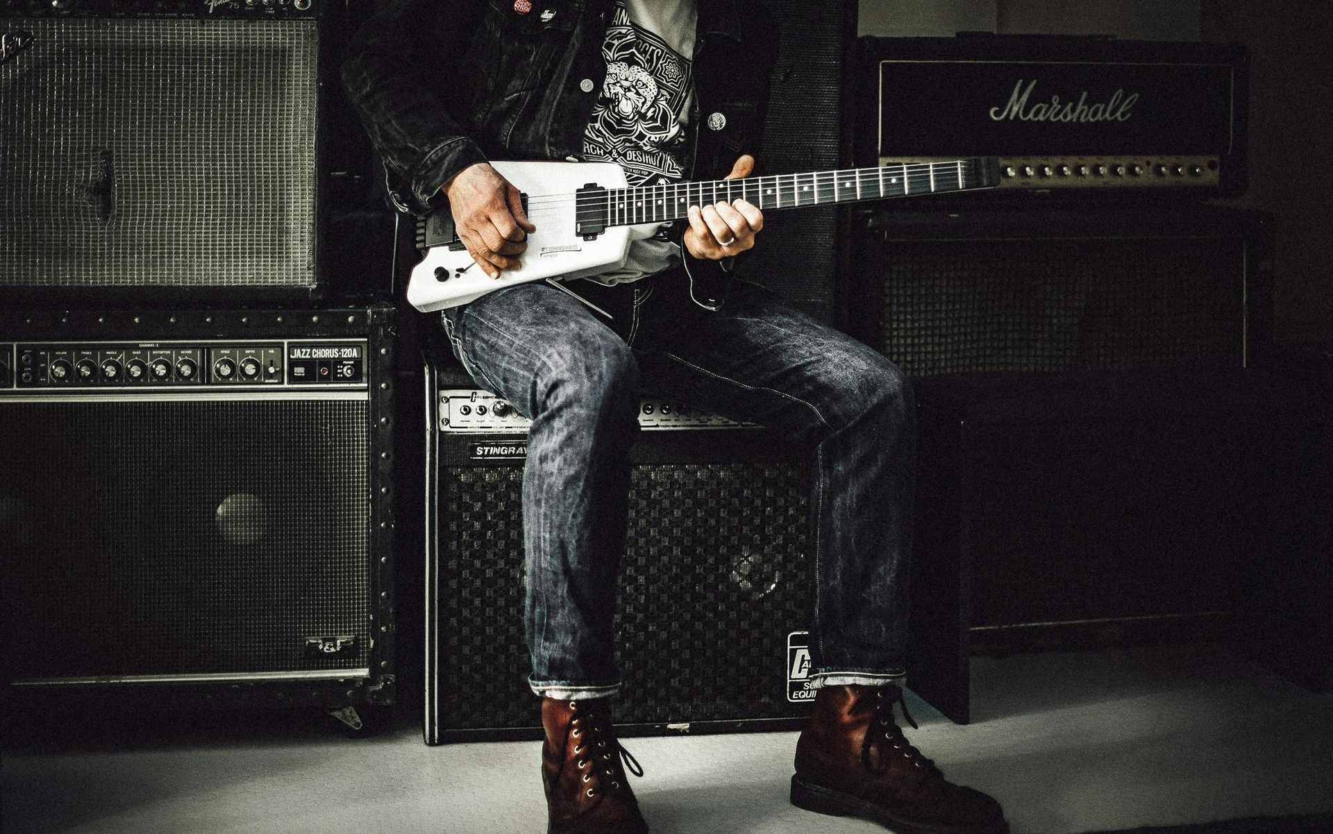 Guitarist playing, seated. Black denim jacket, acid-wash jeans, boots. Amplifiers in background.