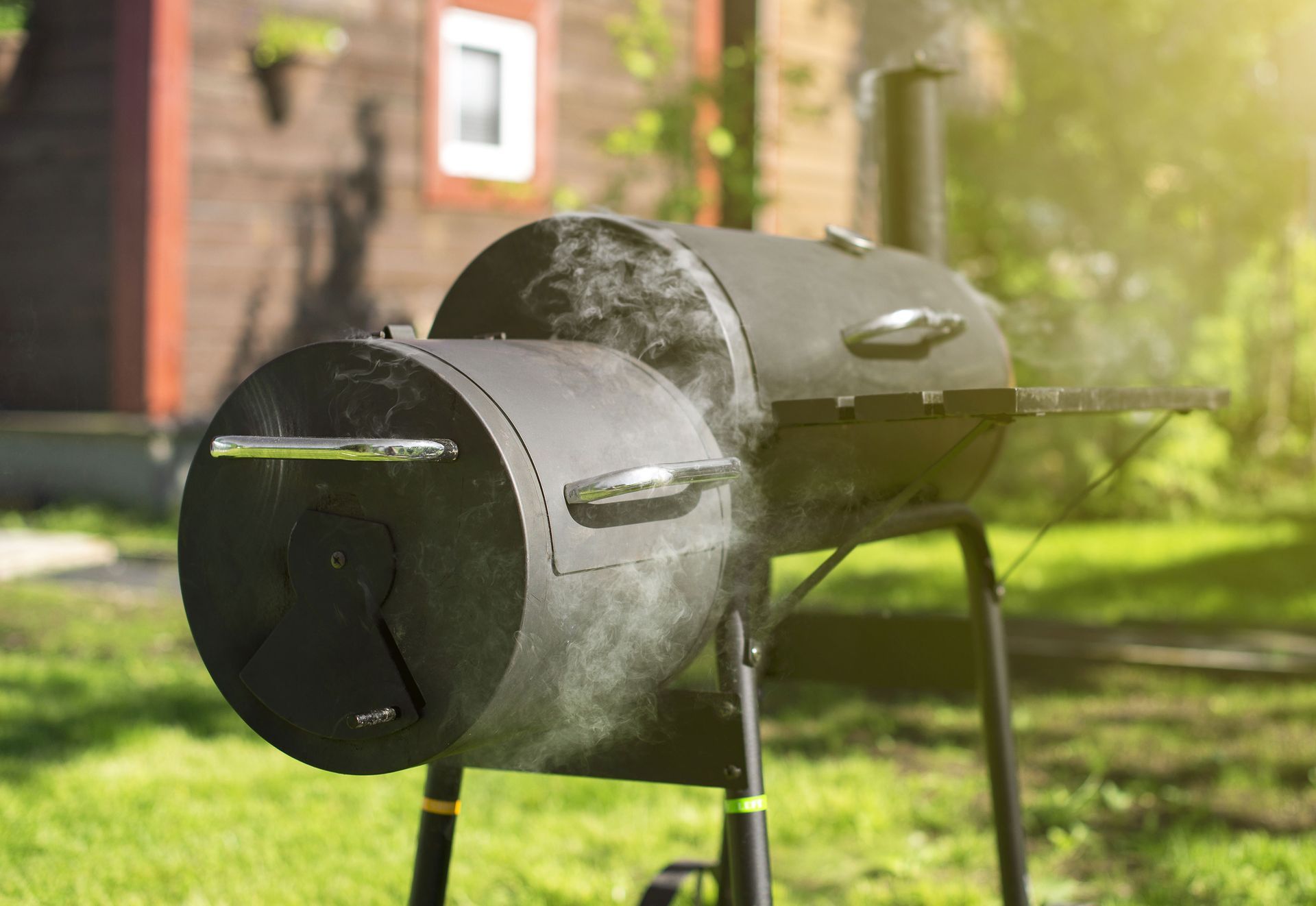 Black smoker grill on a grassy lawn, smoke rising, next to a wood-sided house.