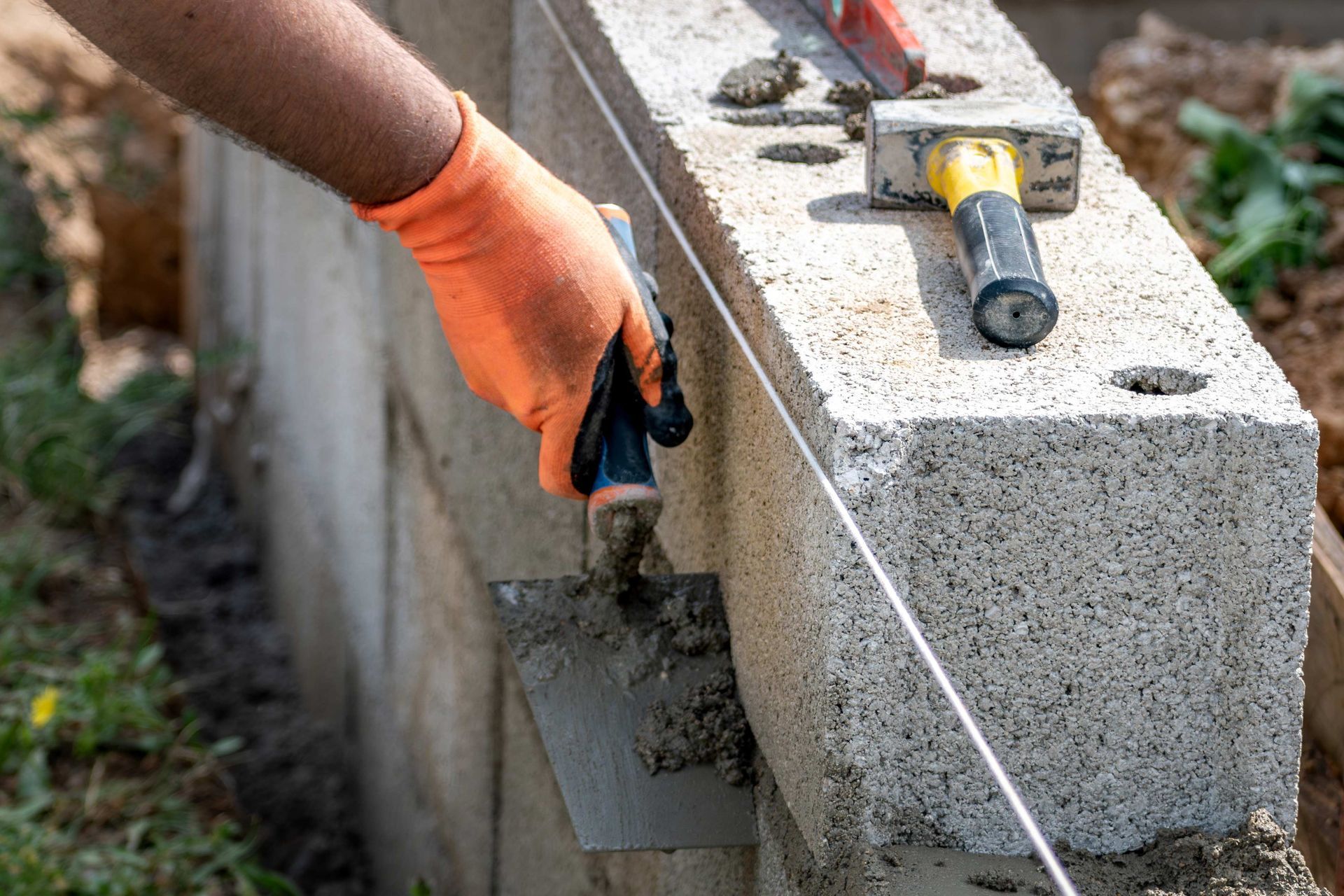 Person in orange glove uses trowel to spread mortar on concrete block wall.