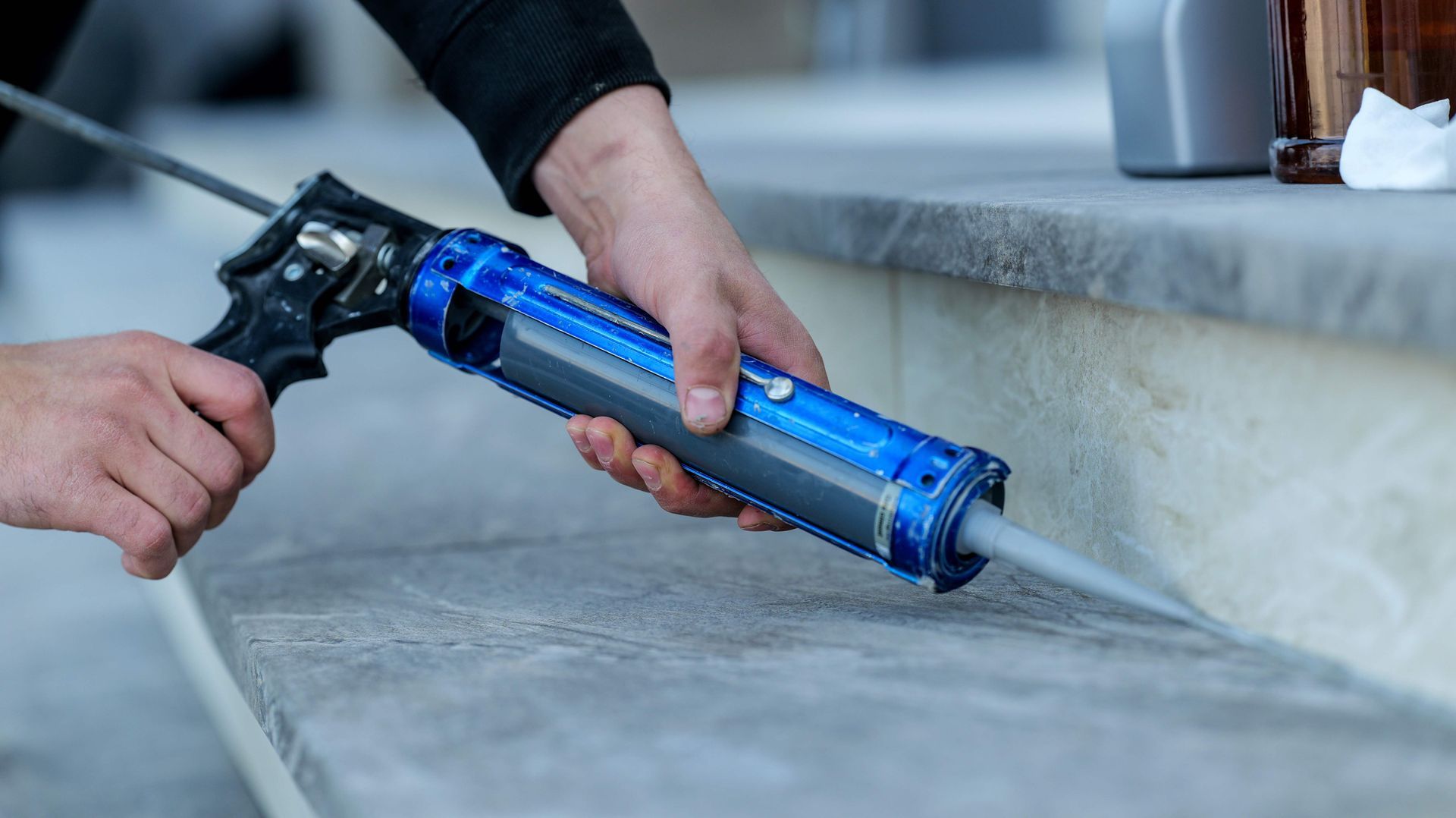 Person using a blue caulk gun to seal a seam on a concrete surface outdoors.