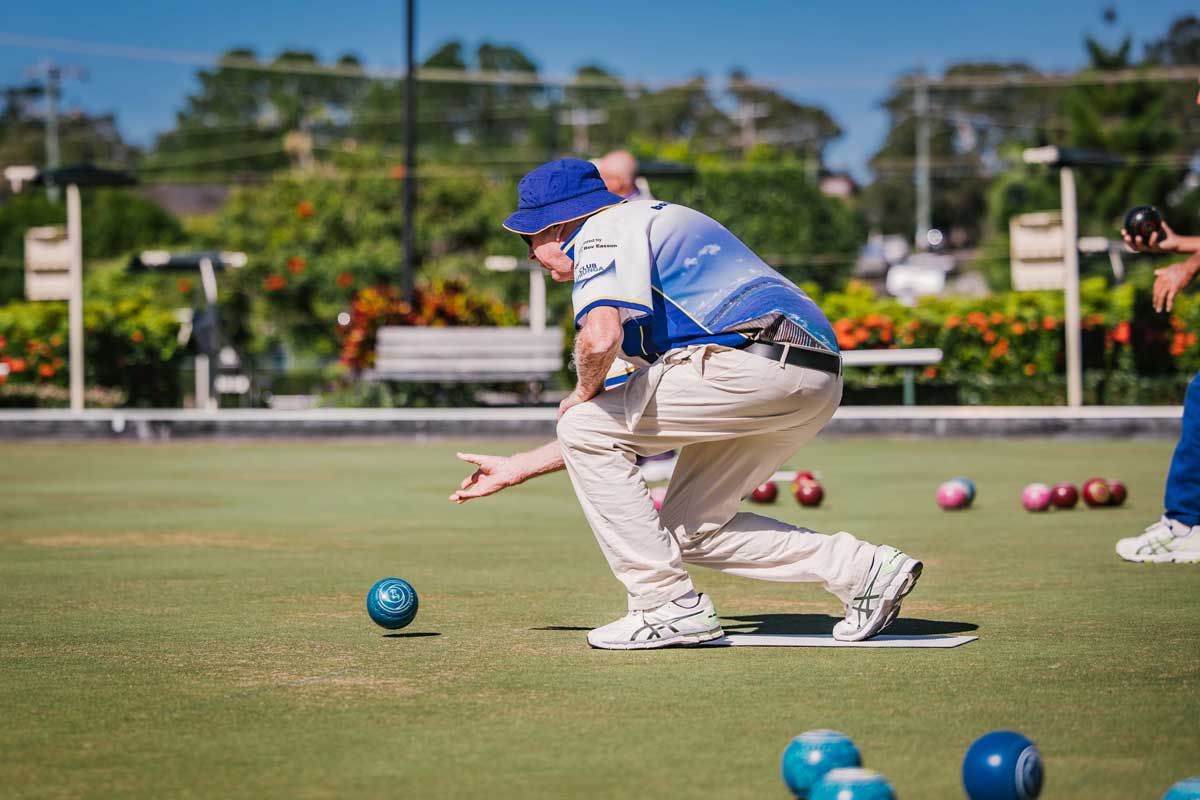 Bowls player in blue and white uniform bowls ball on green lawn toward other bowls. Sunny day.