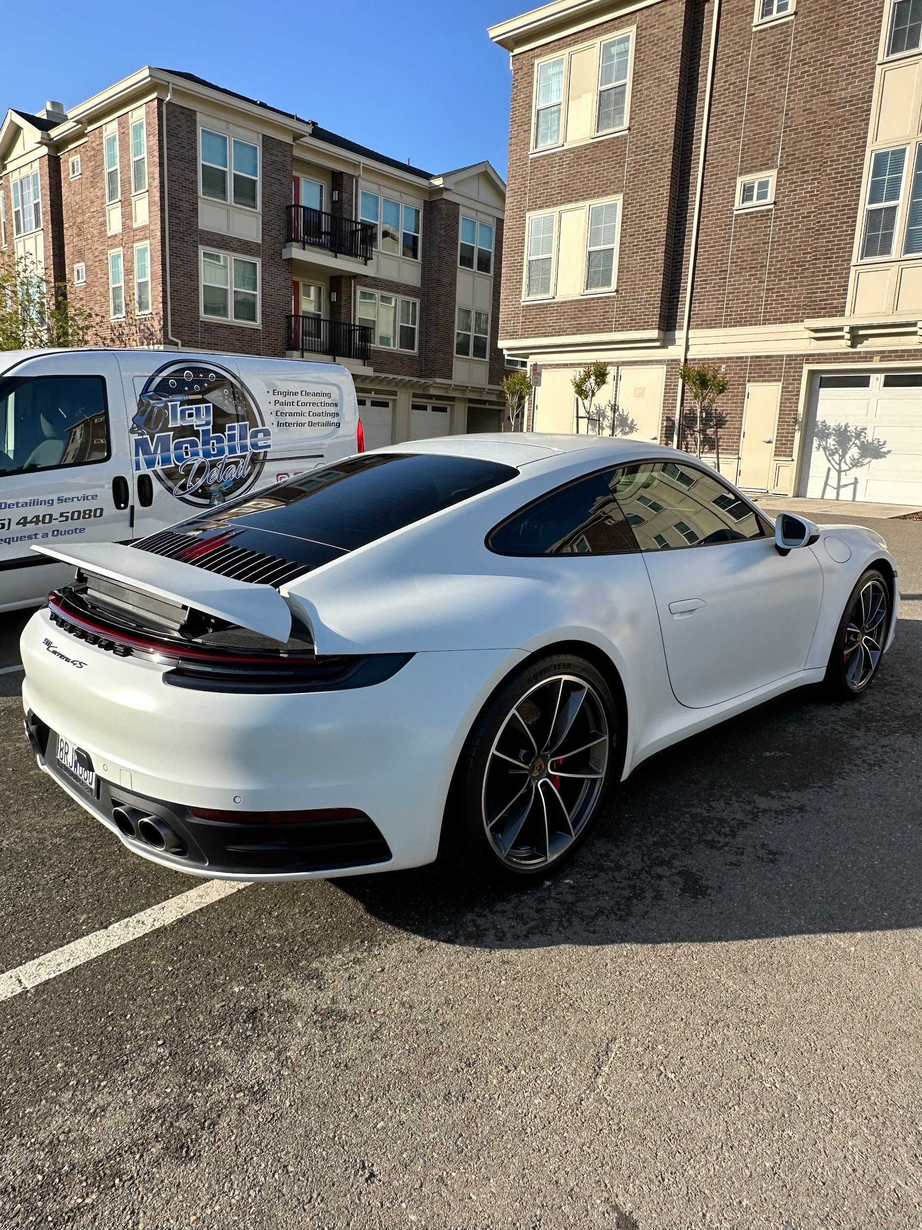 A white porsche 911 is parked in a parking lot in front of a brick building.