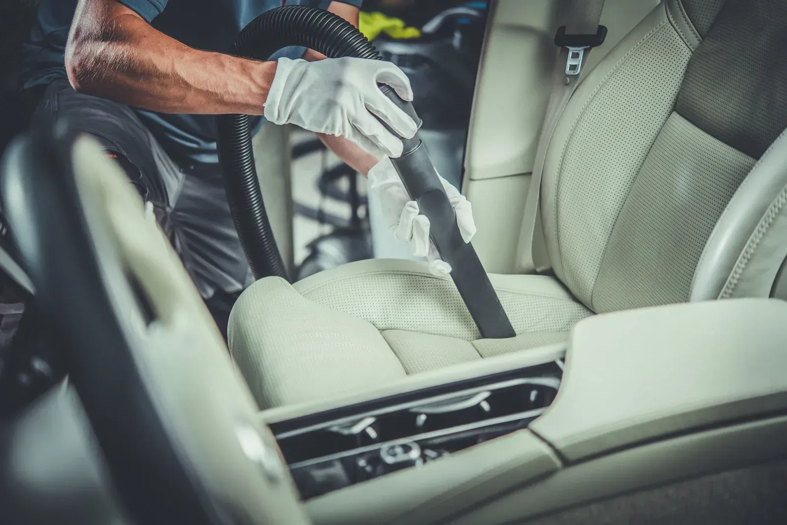 Person vacuuming the cream-colored interior car seat with a vacuum cleaner, wearing white gloves, in a car detailing shop.