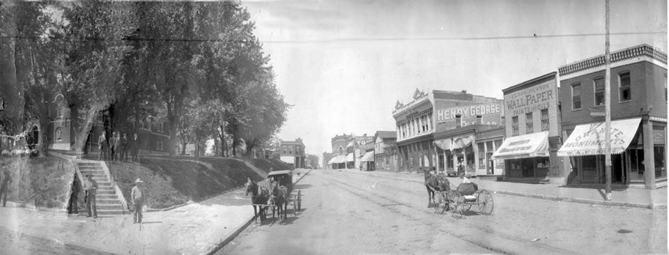 A black and white photo of a city street with horse drawn carriages.