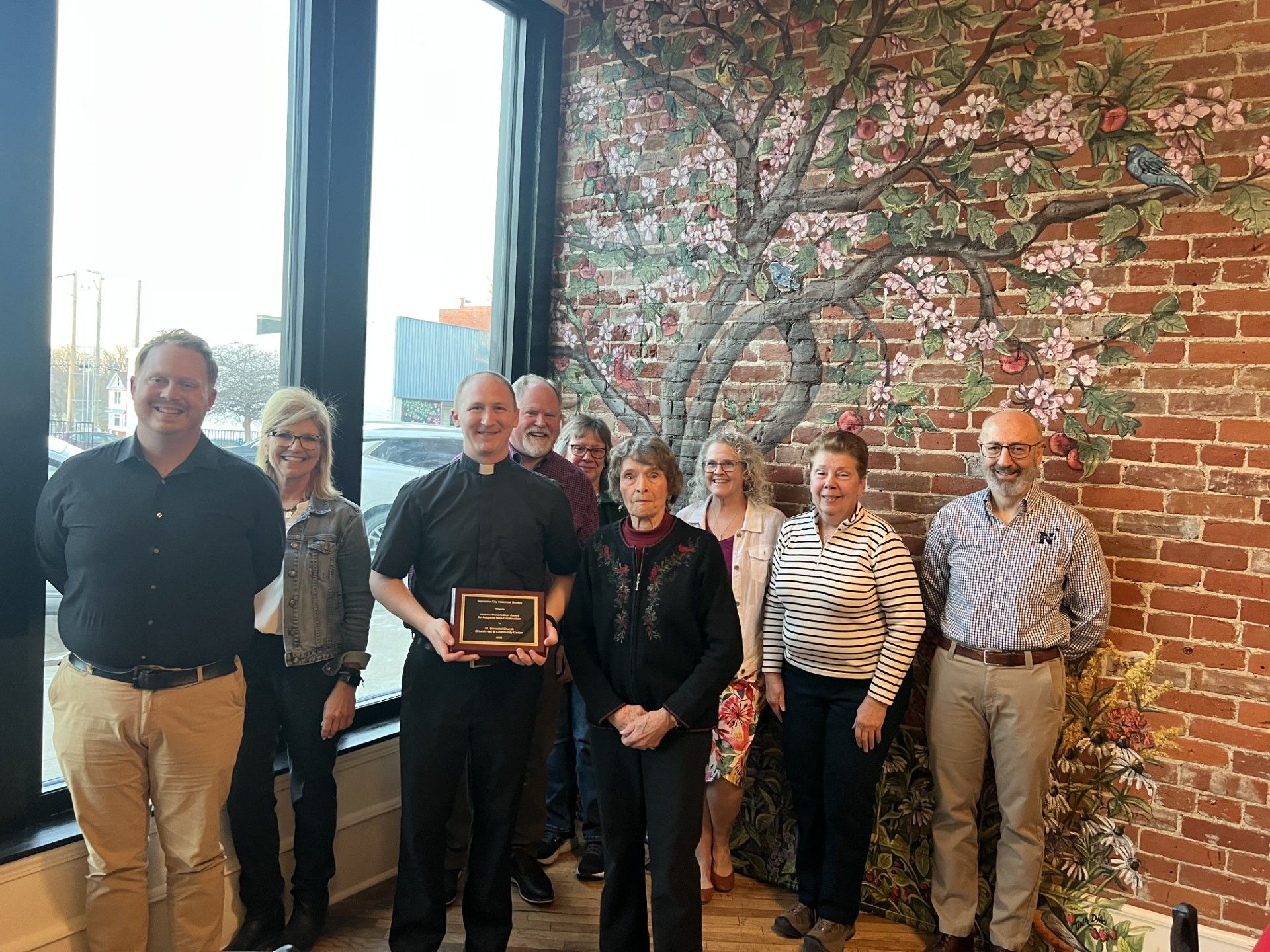 Group of people standing indoors in front of a brick wall, one holding a plaque.