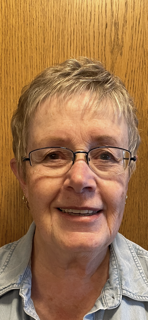 A woman wearing glasses and a name tag is standing in front of a window.