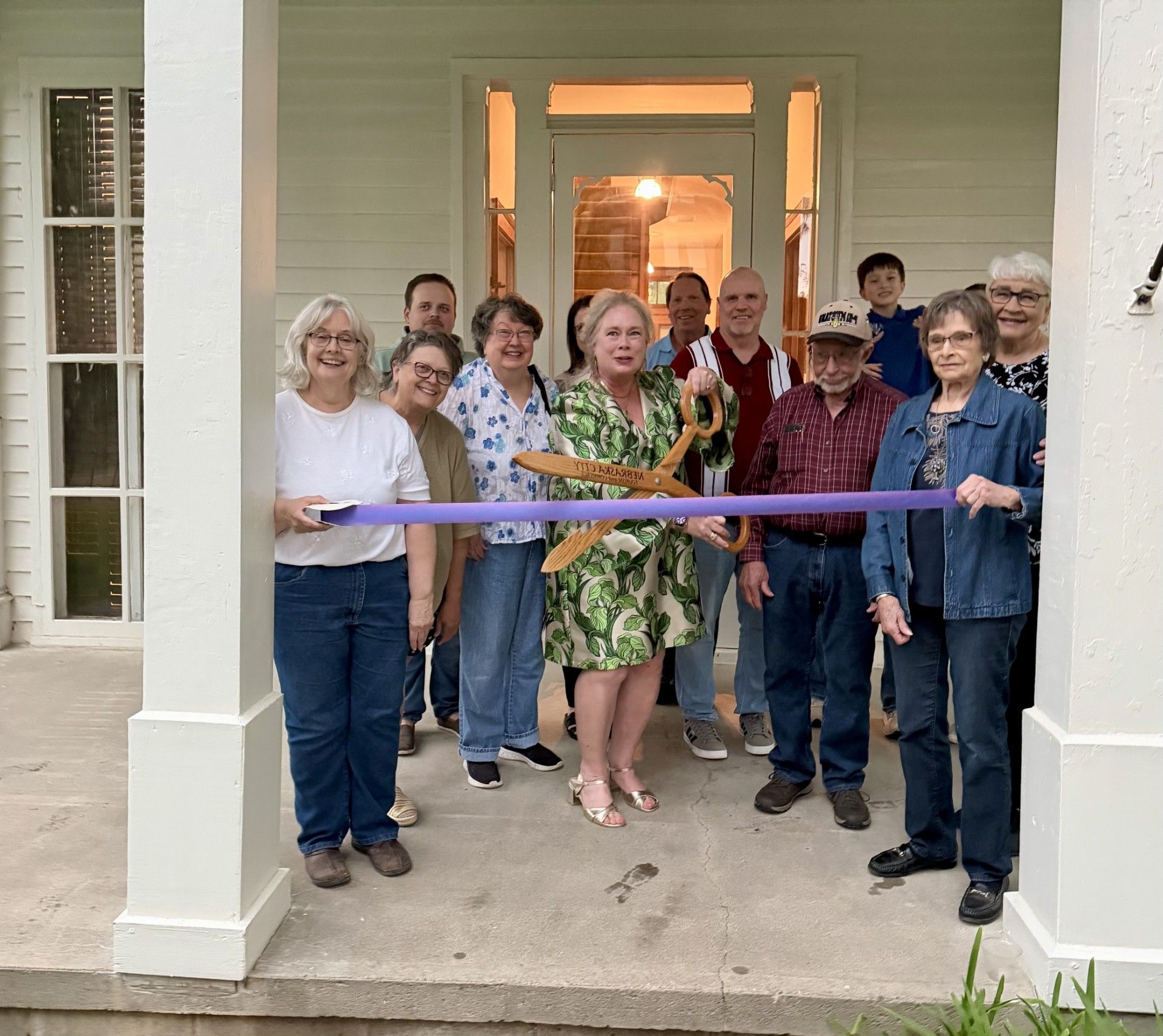 Group of people standing on a porch, smiling and holding a purple ribbon at a ribbon-cutting ceremony