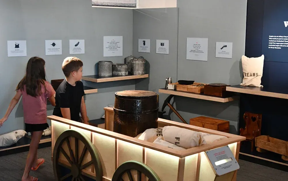 A boy and a girl are standing next to a wooden wagon in a museum.