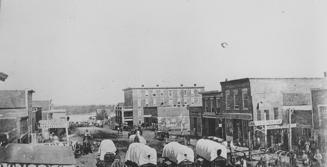 A black and white photo of people riding horse drawn carriages down a street.