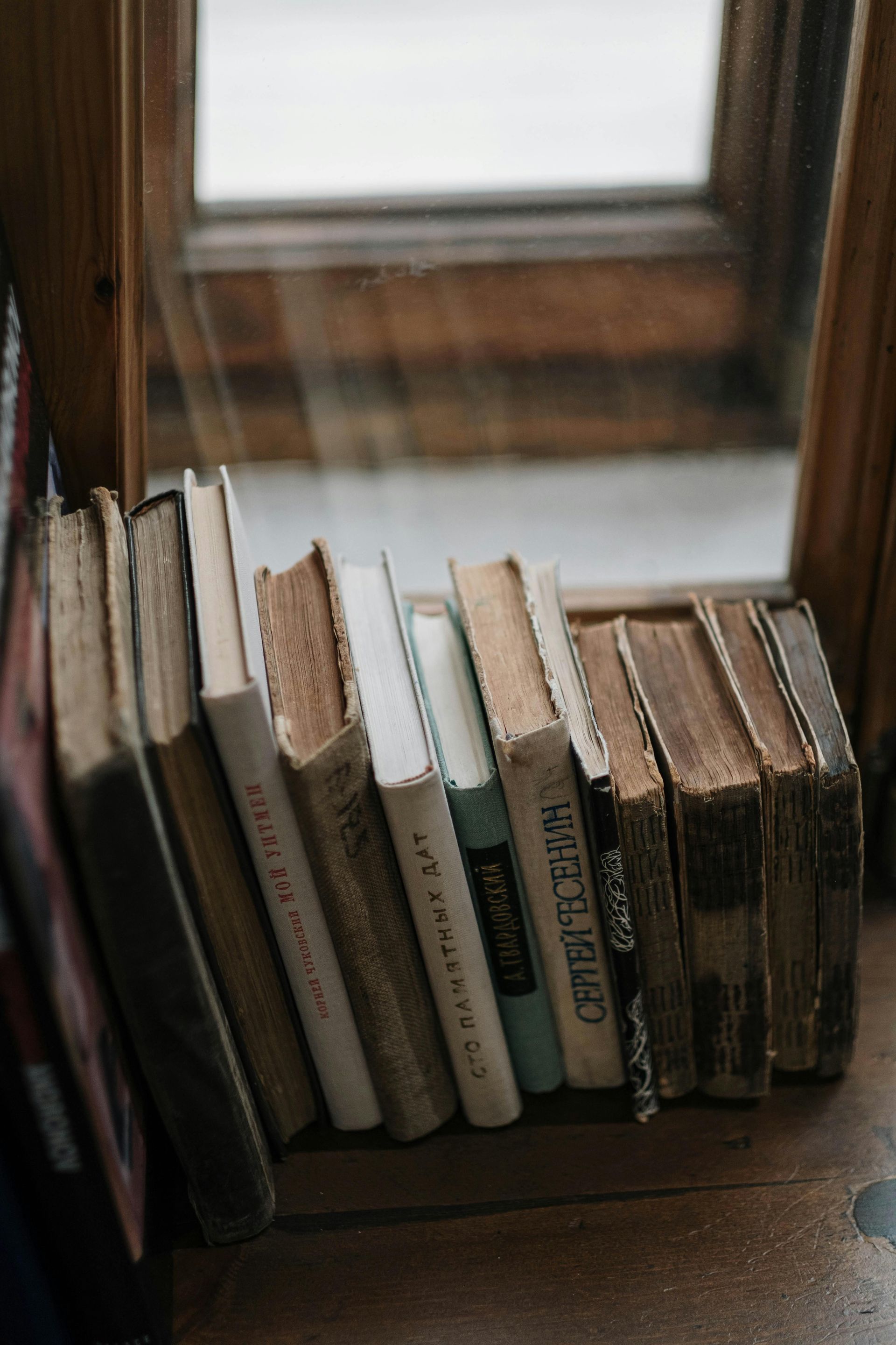 A row of worn books leaning on a wooden windowsill by a window.