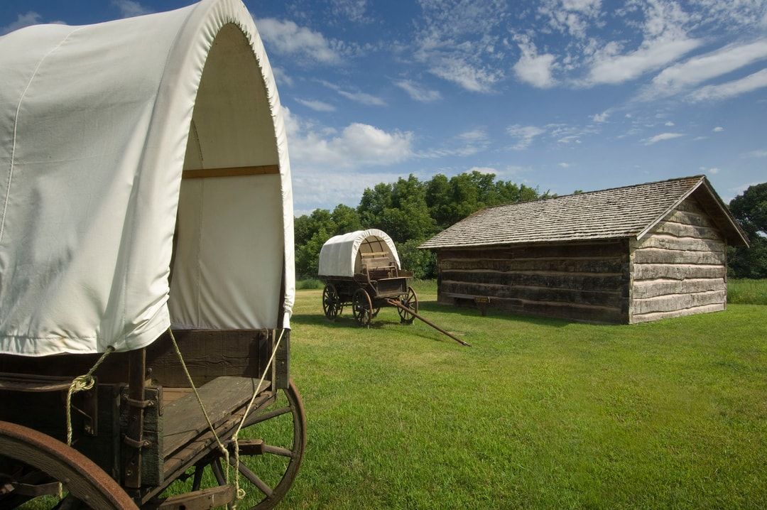 Two covered wagons are parked next to a log cabin in a grassy field.