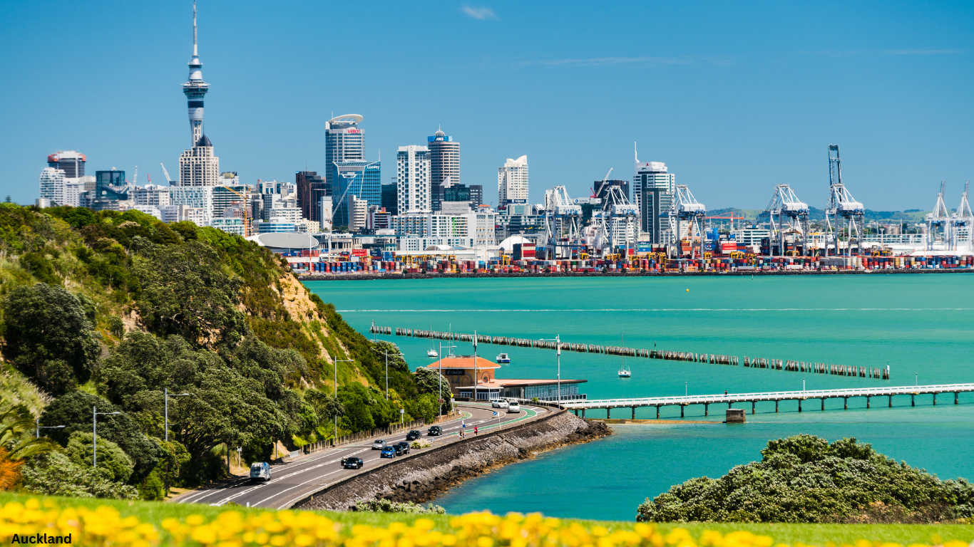 Auckland skyline, including the Sky Tower, visible behind green hills and turquoise harbor.