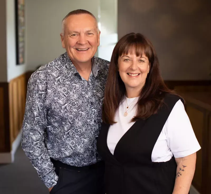 Smiling man and woman standing close together indoors, man in patterned shirt, woman in black vest.