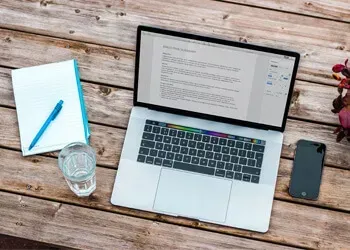 Laptop with document open, next to notebook, pen, phone, and glass of water on wooden table.