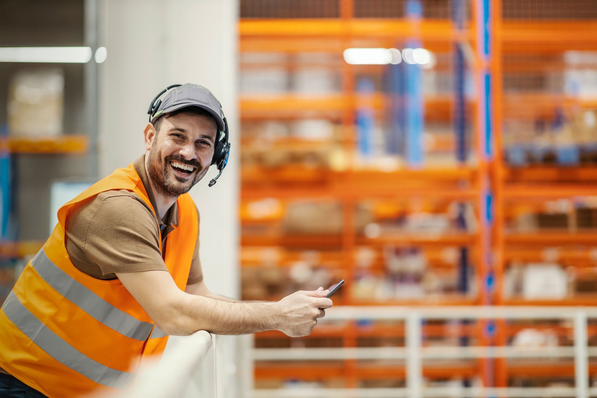 Warehouse worker smiles, leaning on railing, wearing headset and safety vest, holding phone.