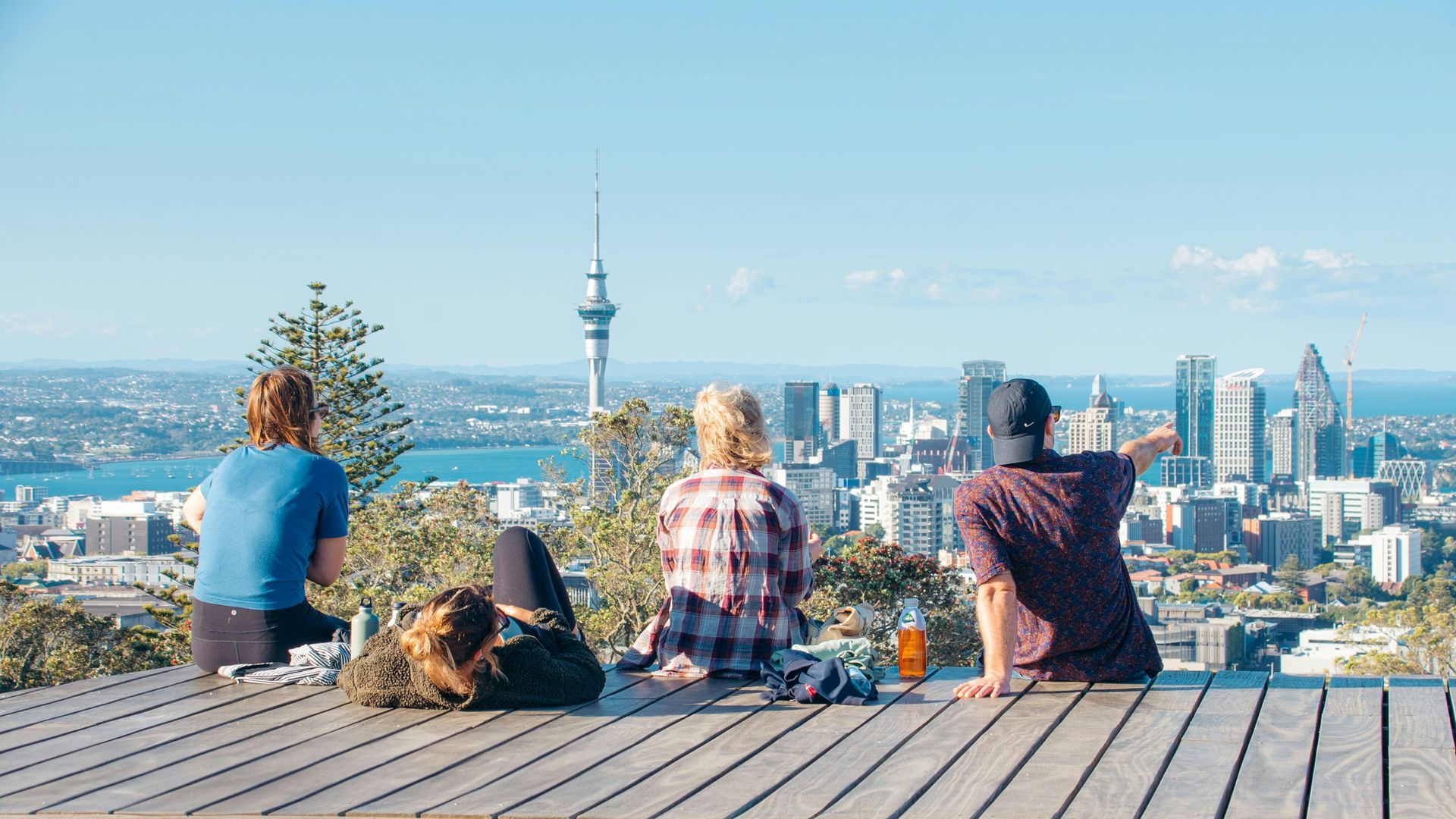Three people sit on a wooden platform overlooking Auckland city, including the Sky Tower, on a sunny day.