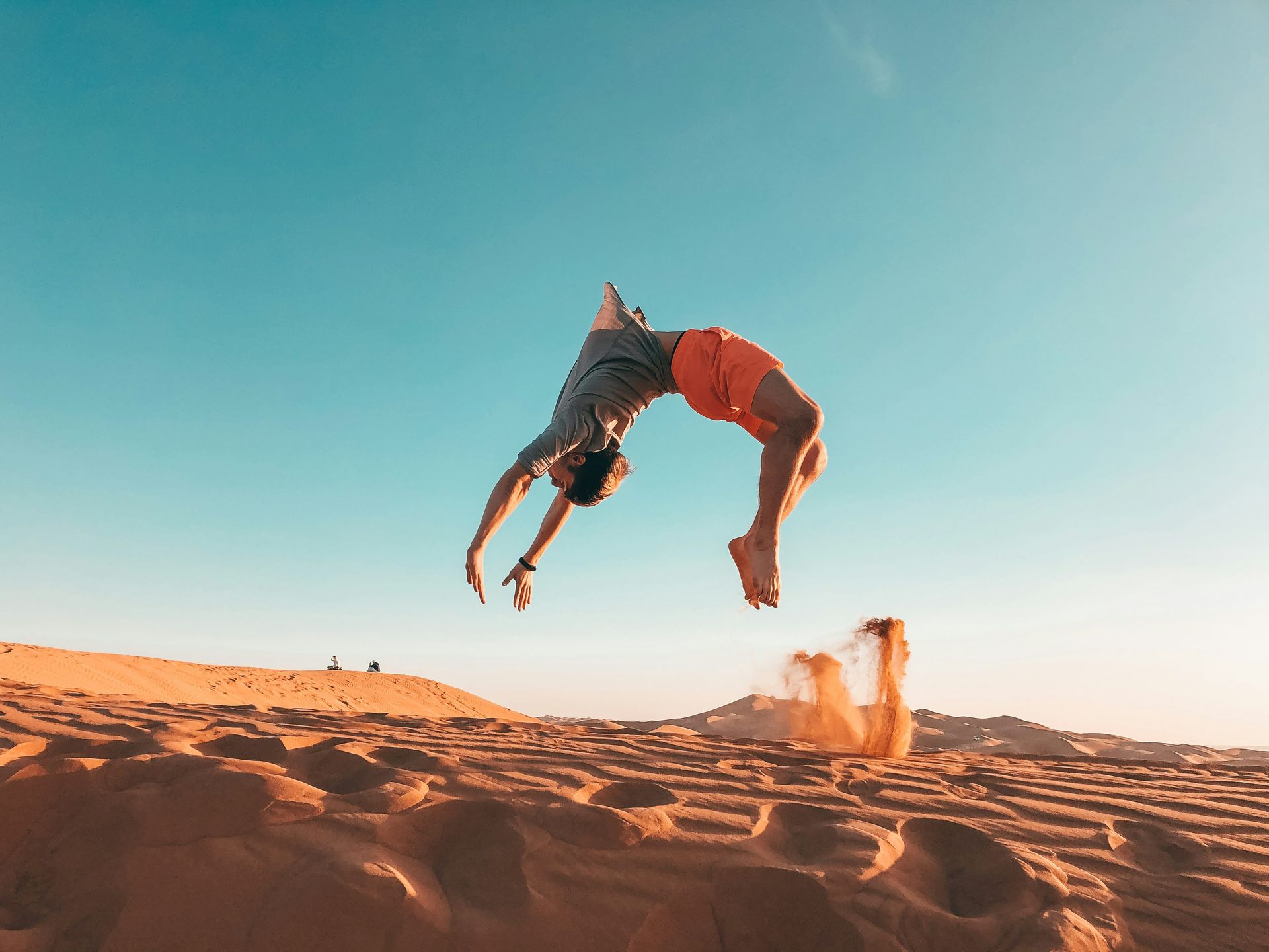 Man doing a backflip in the desert. Orange shorts, sandy ground, blue sky.