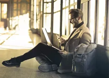 Man in glasses smiles while using laptop and phone, sitting on floor near bag, with window background.