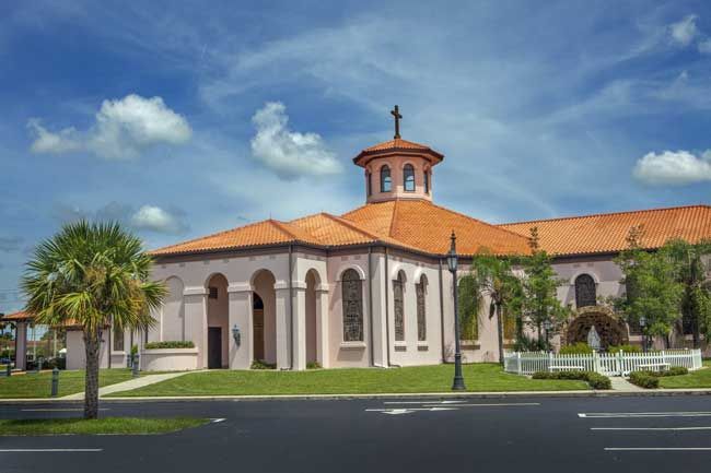 Church with a pink stucco exterior, terracotta roof, and a central dome; palm trees and blue sky background.