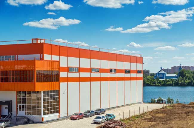 Orange and white industrial building with cars parked in front, blue sky with clouds, near water.