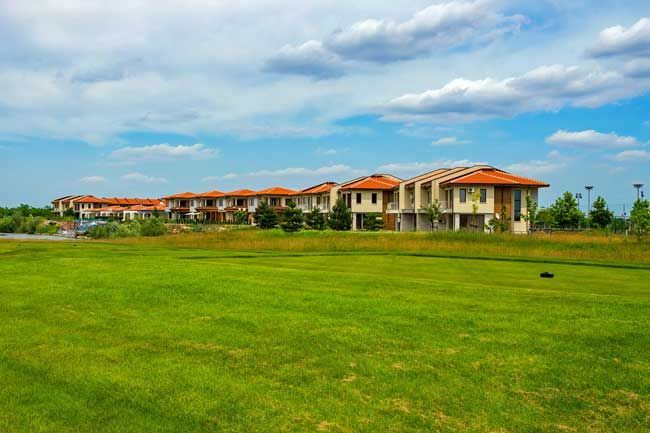 Row of beige houses with orange tile roofs, on a green field under a blue cloudy sky.