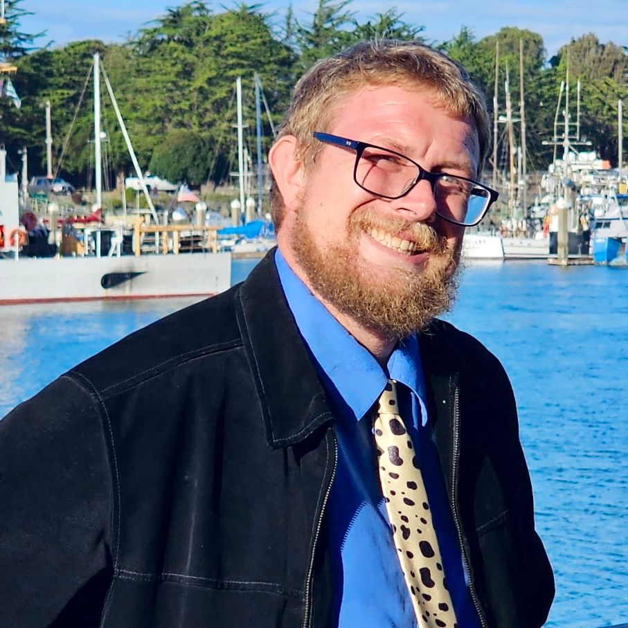Man with glasses and beard smiles at camera, standing in front of a harbor with boats.