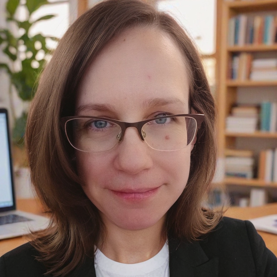 Woman with brown hair and glasses, smiling, in front of a laptop and bookshelf.