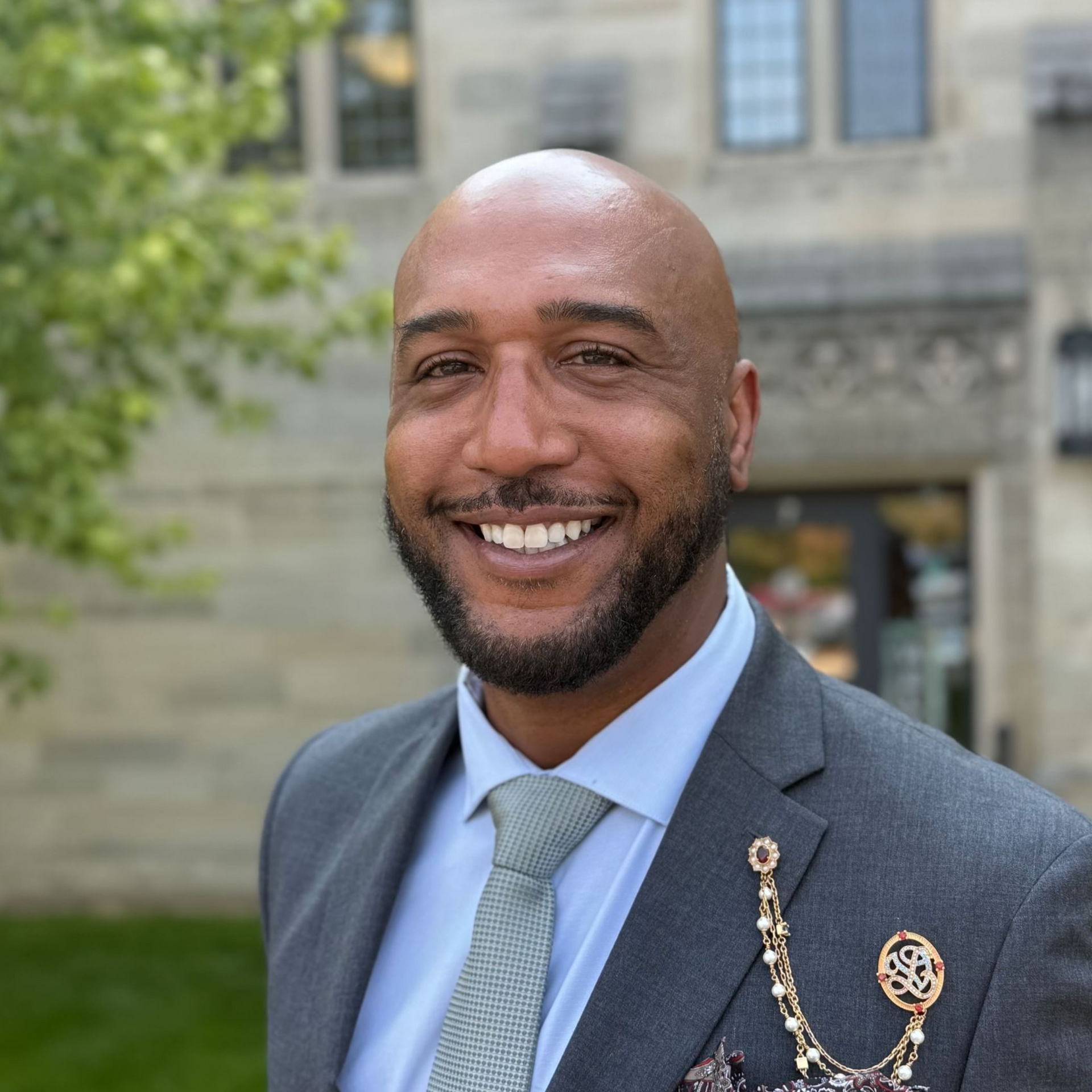 Smiling Black man in a gray suit, tie, and brooch in front of a stone building.