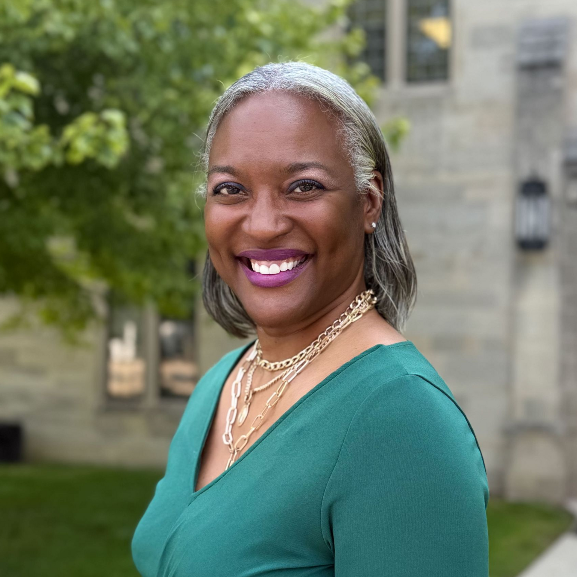 Woman with gray hair smiles, wearing green top and gold necklaces, outdoors near a building.