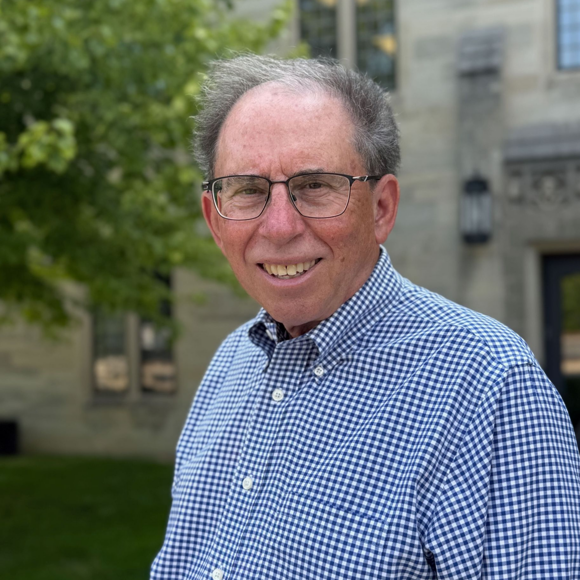 Smiling man with glasses and blue checkered shirt, outside a stone building with trees.