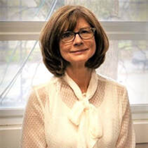 Woman with glasses, brown hair, smiling, wearing a cream-colored blouse, in front of a window.