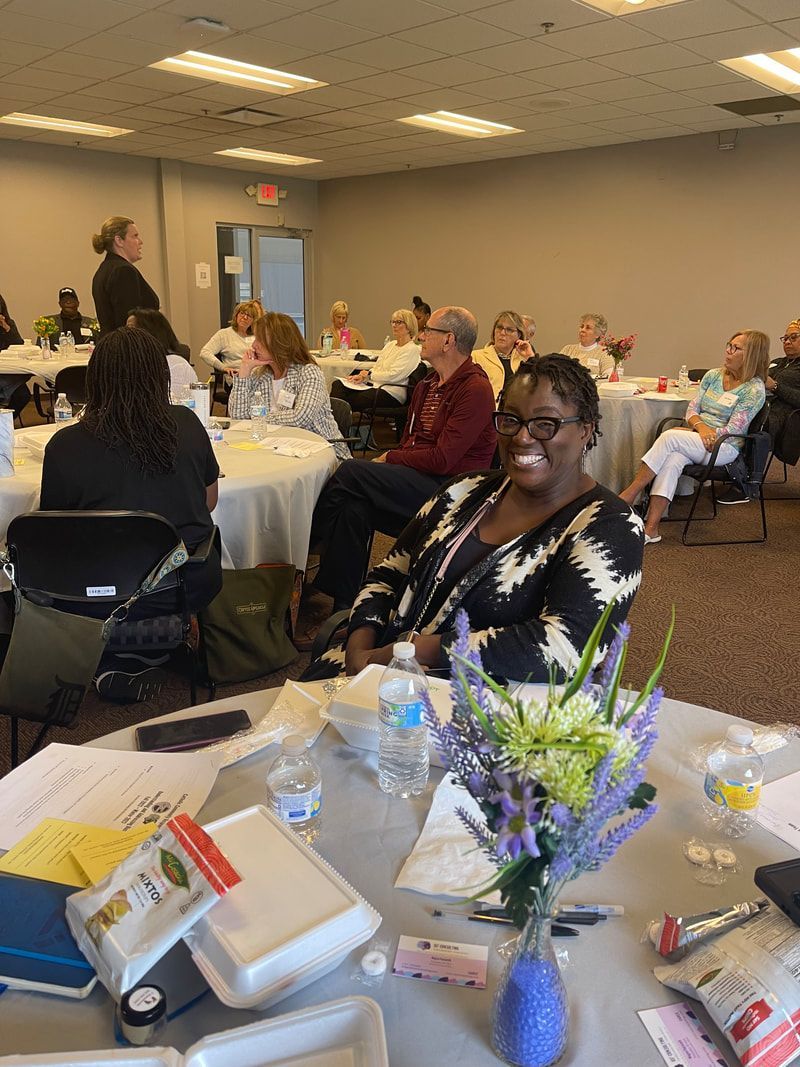 Woman smiling at a meeting with other people seated at round tables.