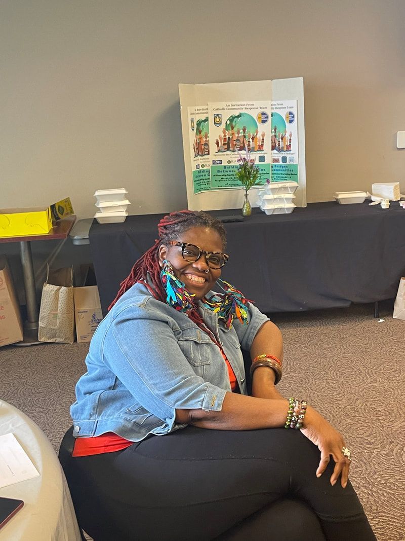 Woman with red braids, denim jacket, smiling, sitting. Table with food and poster in background.