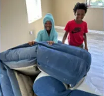 Two young boys smiling while lifting a blue couch in a room with light walls.