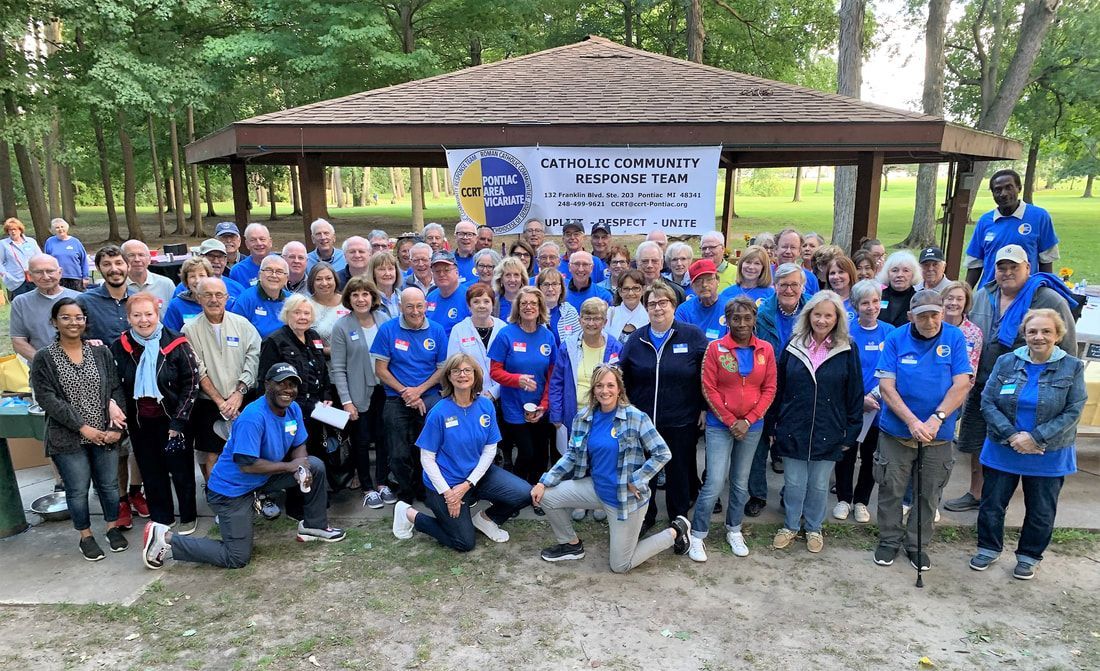 Group of people in blue shirts pose in front of a gazebo, park setting. Banner overhead.