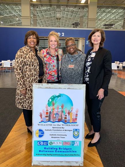 Four women pose next to a sign, “Building Bridges Between Communities” in an event setting.