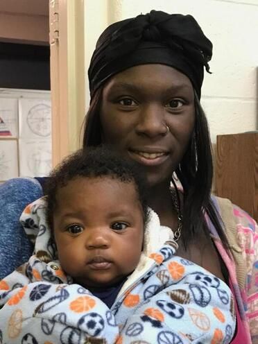 Woman in black headwrap holds baby in patterned blue hoodie, both looking at camera.