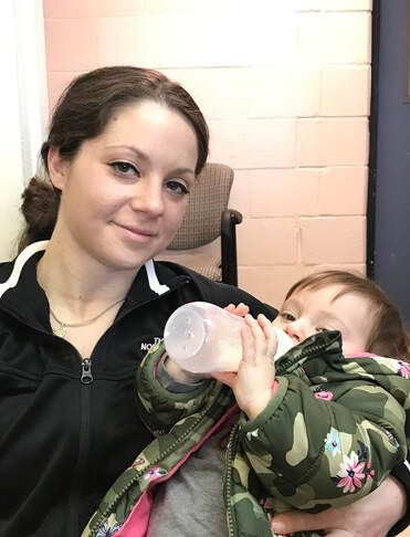 Woman smiles, holds baby drinking from bottle. Both indoors, against light-colored wall. Baby wears camo jacket.