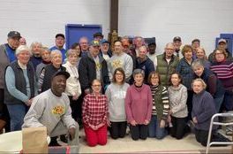 Group of diverse people smiling, indoors. Some kneel. White, blue walls in background.