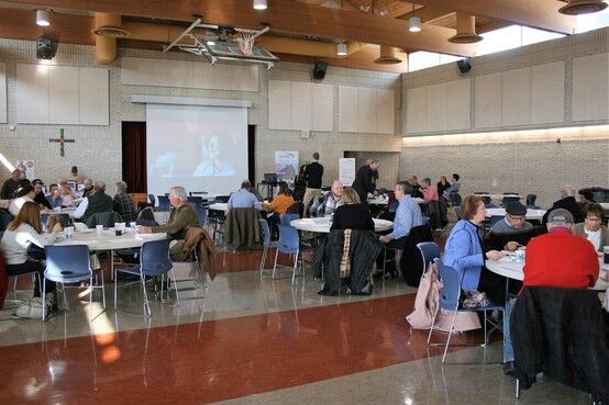 Large meeting room, people seated at round tables, watching a presentation on a screen.