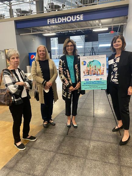 Four women stand near a sign in a fieldhouse. They are all smiling.