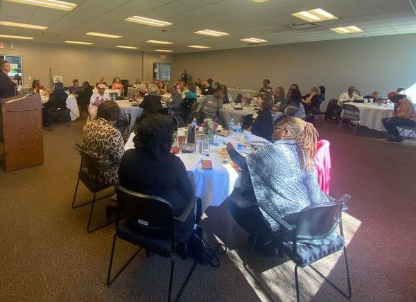 People seated at round tables in a large room. A speaker at a podium is visible on the left.