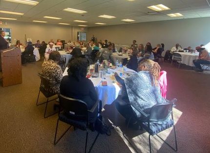 People seated at round tables in a large room. A speaker at a podium is visible on the left.
