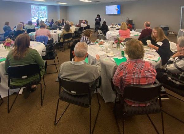 People seated at tables in a room, listening to a presenter. Some tables have floral centerpieces.