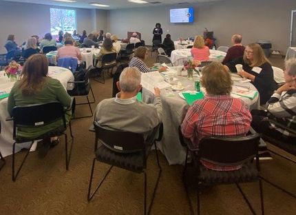 People seated at tables in a room, listening to a presenter. Some tables have floral centerpieces.